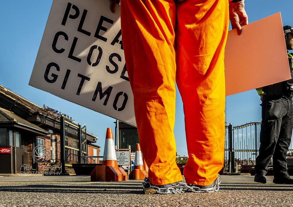 Protesters legs wearing orange jump suit with changes around ankle holding a placanard saying please close GITMO