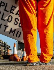 Protesters legs wearing orange jump suit with changes around ankle holding a placanard saying please close GITMO