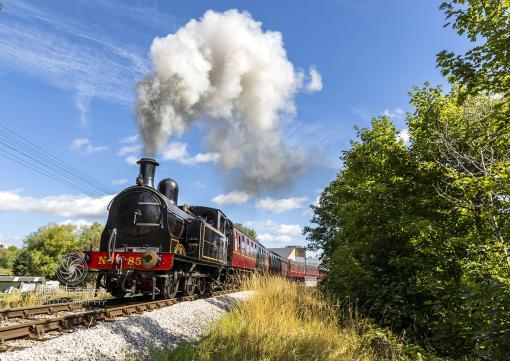 Steam train Taff Vale Railways no.85 with white steam under blue sky with green trees at the right side of the frame