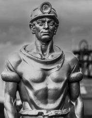 Black and white statue of a miner wearing helmet and lamp adorned with coal not dole stickers with pit headgear in background